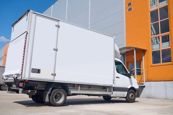 Car, warehouse. White cargo covered vehicle standing outdoors in parking lot near warehouse on fine day